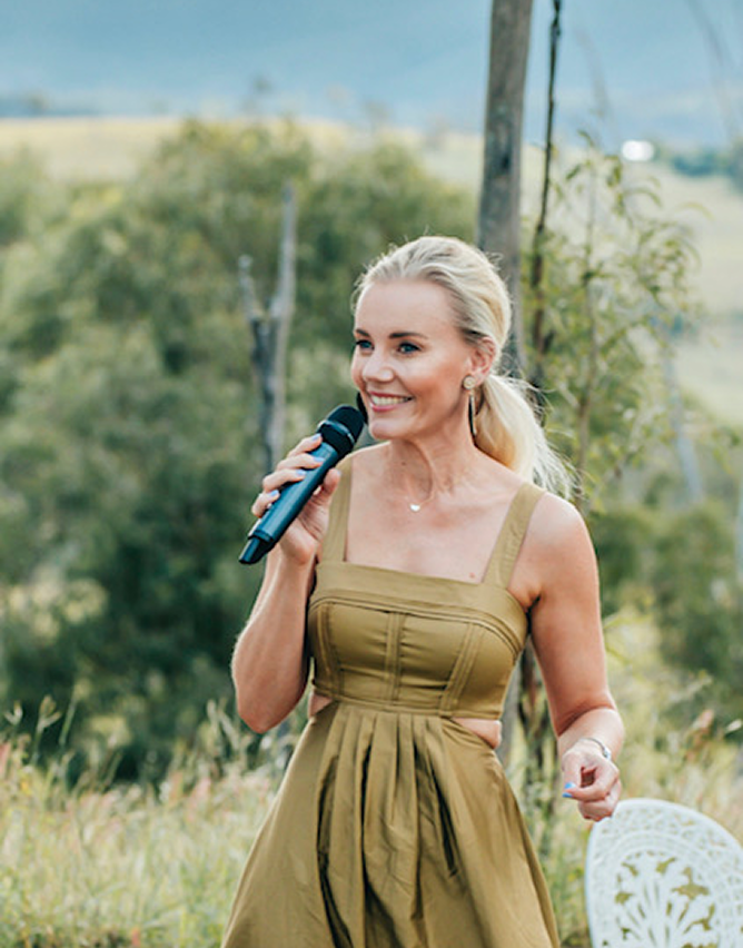 A woman with blonde hair in a ponytail, wearing a khaki-colored dress, holding a microphone, outdoors with green trees in the background.
