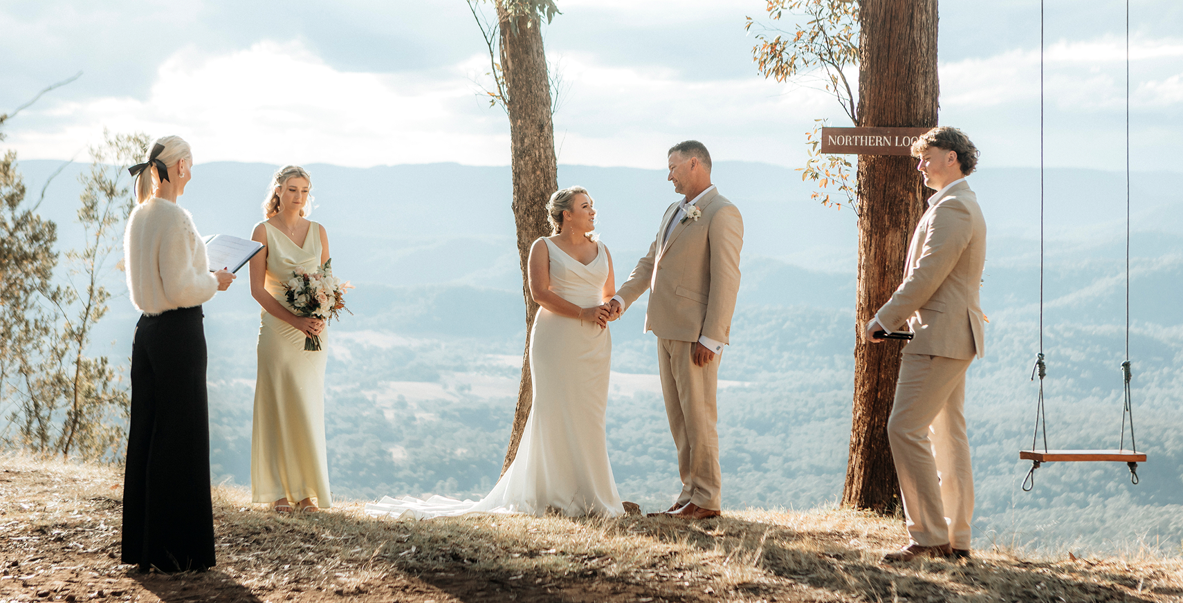 A wedding ceremony taking place outdoors on a hillside overlooking mountains, with a bride and groom holding hands and exchanging vows, officiant reading from a book, and a bridesmaid holding flowers. A sign reading 'Northern Look' is attached to a tree, and a swing hangs from another tree.