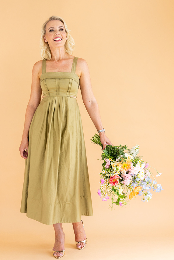 A smiling woman in a light green dress holding a bouquet of colorful flowers against a light peach background.