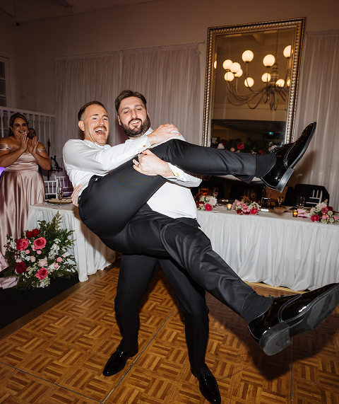 Two men in formal wear dancing joyfully at a wedding reception, with one lifting the other off the ground. A woman in a pink dress and other guests are visible in the background, along with decorated tables and a large mirror reflecting a chandelier.
