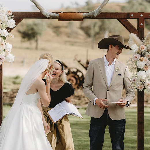 Bride and groom at an outdoor wedding ceremony, with an officiant, under a rustic wooden arch decorated with flowers, in a rural setting.