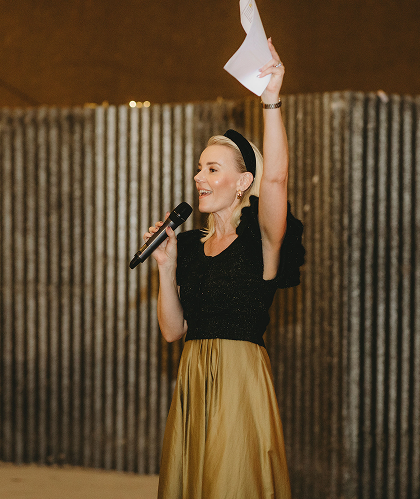 Woman speaking into a microphone, holding a paper in the air, in front of a wooden backdrop.