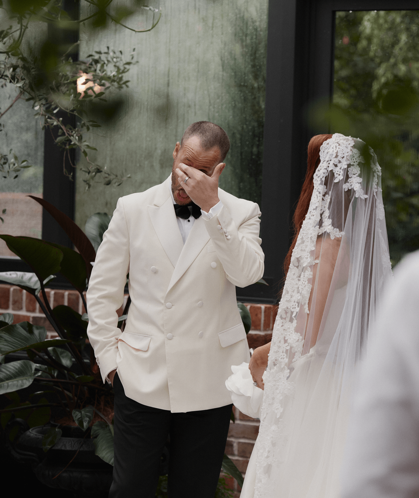 A man in a white tuxedo jacket holding his face with one hand, appearing emotional, next to a woman in a wedding dress with a lace veil during a wedding ceremony.