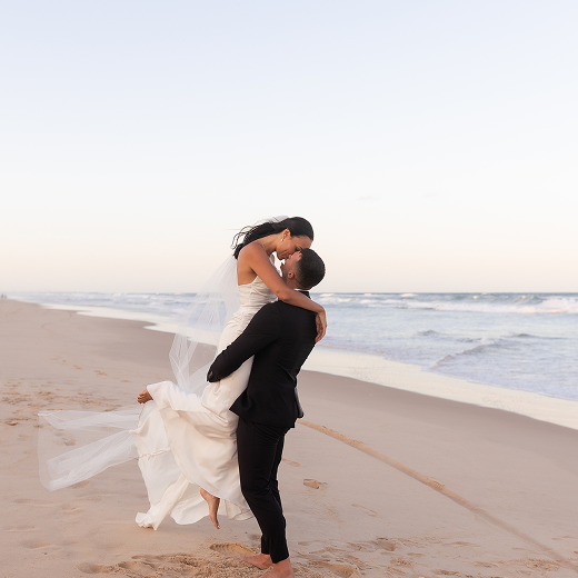 A bride and groom on the beach, with the groom lifting the bride while they kiss, during sunset or early evening.