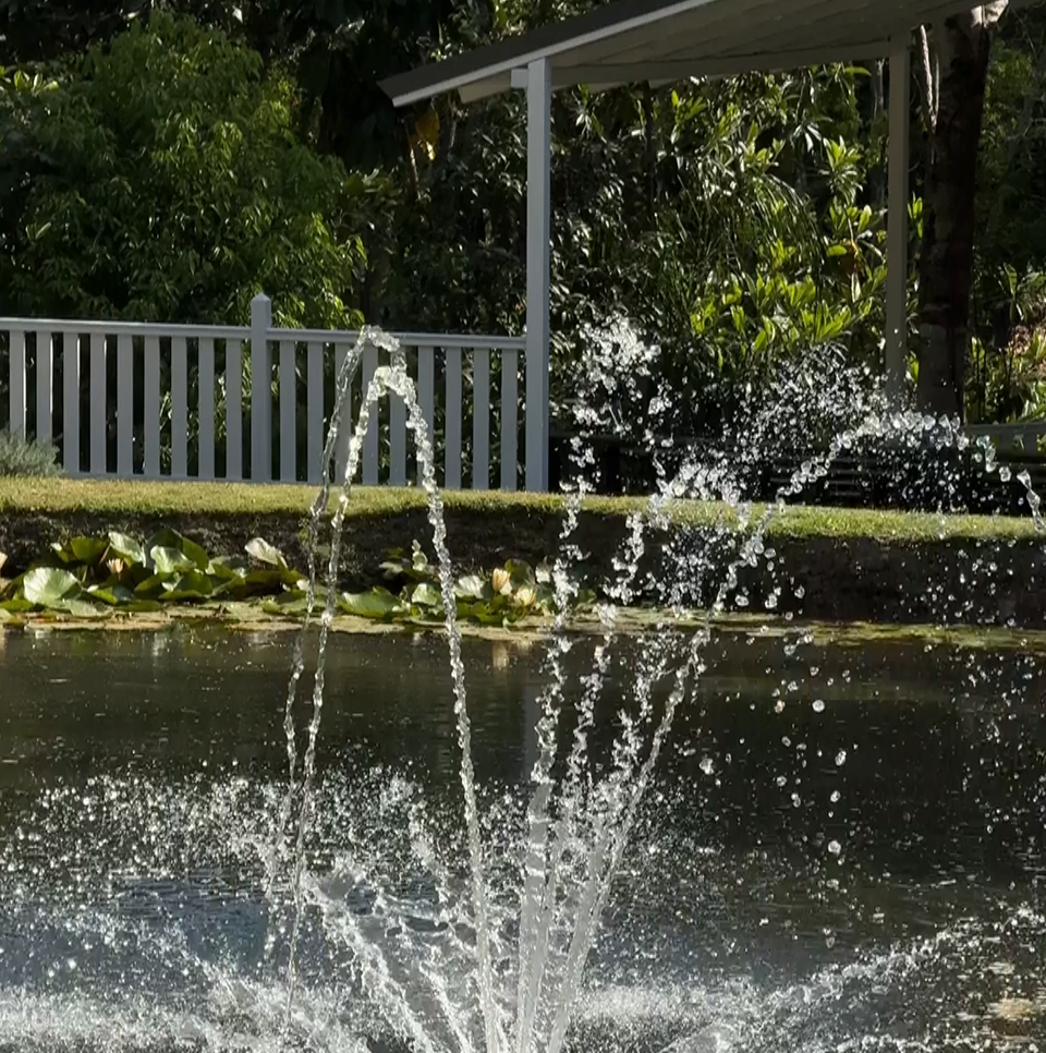 A water fountain spraying water into the air in a pond or small lake, with greenery and trees in the background, and a wooden structure with a white railing in the scene.