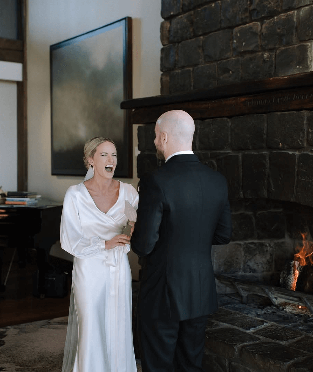 A woman in a white dress is laughing with a man in a black suit by a fireplace.