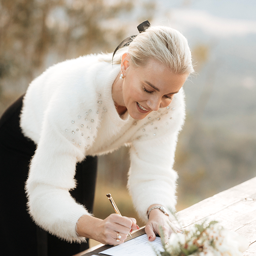 Woman with blonde hair in a ponytail, wearing a white sweater with pearl accents, signing a book outdoors with a smile.