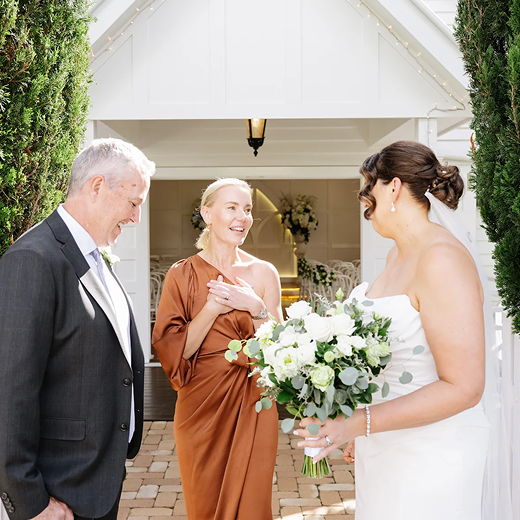 Bride in white wedding dress holding a bouquet of white flowers and greenery, talking with a woman in a brown dress, with an older man in a dark suit standing nearby in front of a small white chapel with trees on each side.