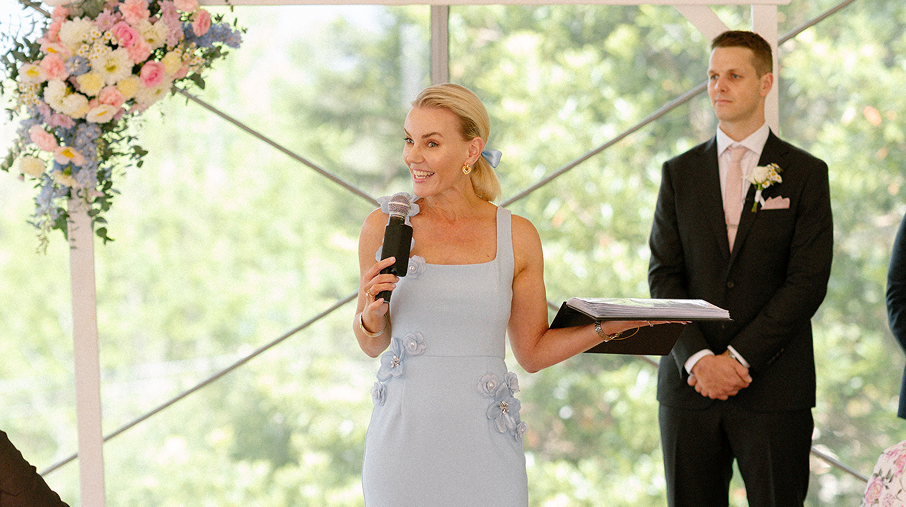 A woman in a light blue dress, holding a microphone and a black folder, speaking at a wedding ceremony, with a man in a black suit and pink tie standing behind her.