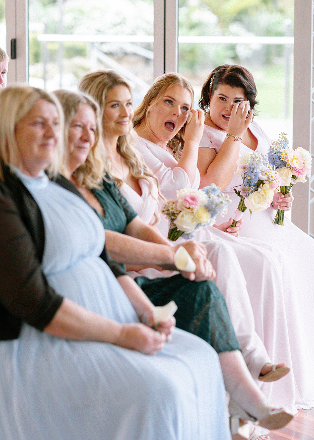 Women sitting in a row at a wedding or similar event, some holding bouquets of flowers, with expressions of joy and emotion.