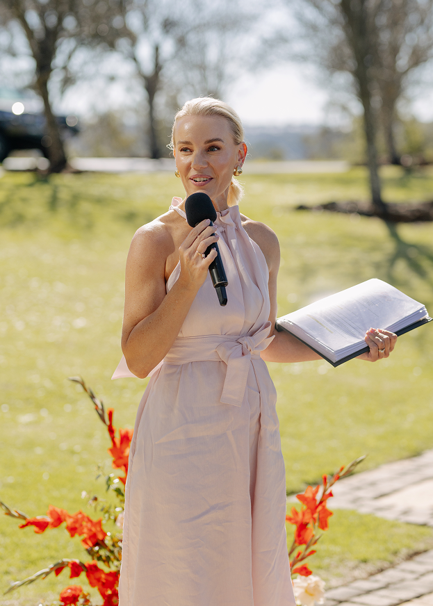 A woman in a light pink dress holding a microphone and a book outdoors during daytime, with trees and flowers in the background.