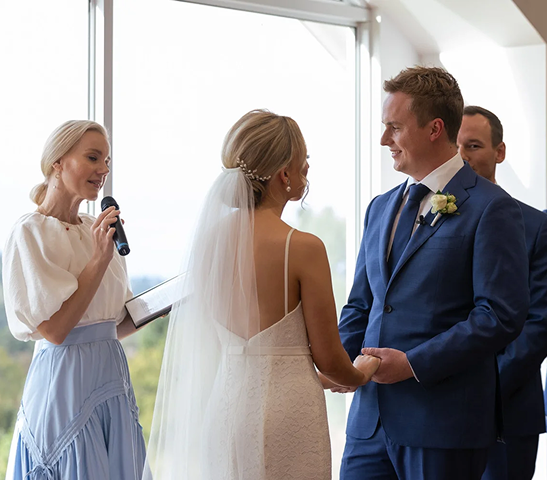 Bride and groom exchanging vows during wedding ceremony indoors, officiant speaking, and a man standing behind the groom.