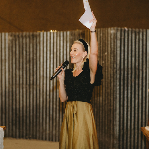 A woman speaking into a microphone while holding a piece of paper, gesturing with her other hand, standing in front of a corrugated metal background, wearing a black top and a gold skirt.