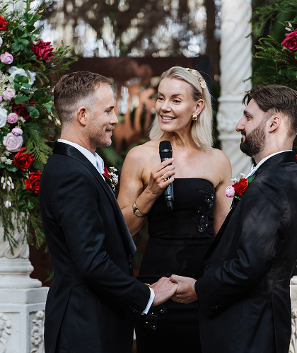 Two men in tuxedos holding hands during a wedding ceremony, with a woman officiant in a black dress and blonde hair, standing between them and smiling, in an outdoor garden setting.