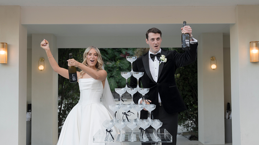 Bride and groom celebrating at their wedding, pouring champagne into a tower of glasses.