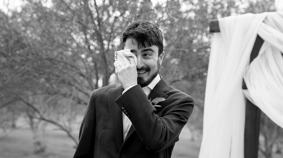 A man in a tuxedo smiling and wiping away a tear during an outdoor wedding or special event.