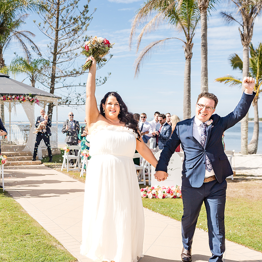 A newlywed couple celebrating outdoors, walking hand-in-hand by a lakeside, with palm trees and guests in the background.