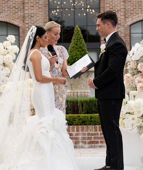 Bride and groom exchanging vows during outdoor wedding ceremony with officiant reading from a book, surrounded by floral arrangements.