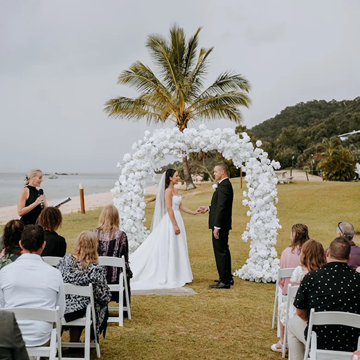 A wedding ceremony outdoors under a palm tree, with the bride and groom holding hands beneath a flower arch, surrounded by seated guests.
