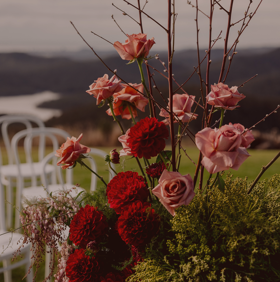 A floral arrangement with pink roses, red dahlias, and other greenery, set outdoors with white chairs and a scenic landscape in the background.