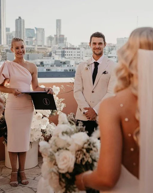 A wedding ceremony on a rooftop with city skyline in the background. The bride, in a wedding dress holding a bouquet, faces the officiant and the groom. The officiant, a woman in a pink dress, holds a folder. The groom, in a white suit with black tie, stands smiling.