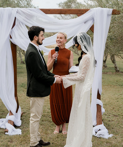 A couple getting married outdoors under a wooden arch draped with white fabric, with an officiant standing behind them.