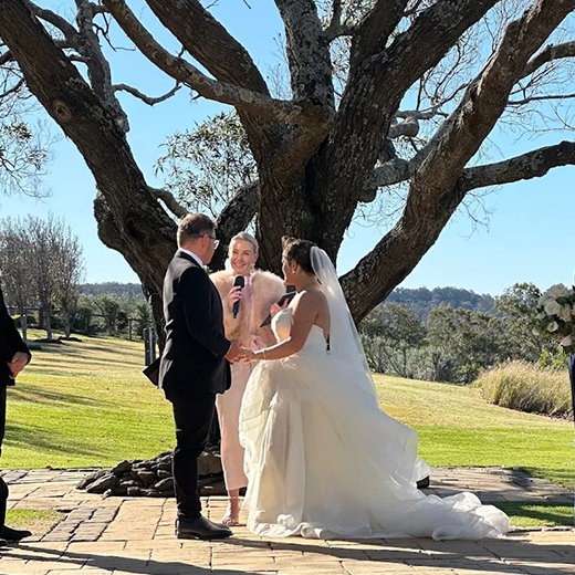 A wedding ceremony outdoors under a large tree, with a bride and groom holding hands and an officiant speaking into a microphone. The bride is wearing a white wedding gown with a train and veil, and the groom is in a black tuxedo. The background features a grassy landscape and clear blue sky.