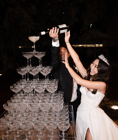 A couple in wedding attire pouring champagne into a tower of glasses at a celebration.