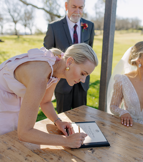 A woman in a pink dress signs a document at a wedding ceremony outdoor under a tent, with a man in a suit and a woman in a wedding dress nearby.