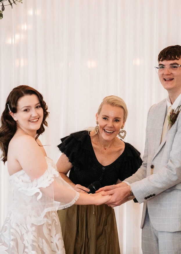 A woman holding hands with a man during a wedding ceremony, smiling in front of a white curtain, with a joyful woman officiant in the middle.