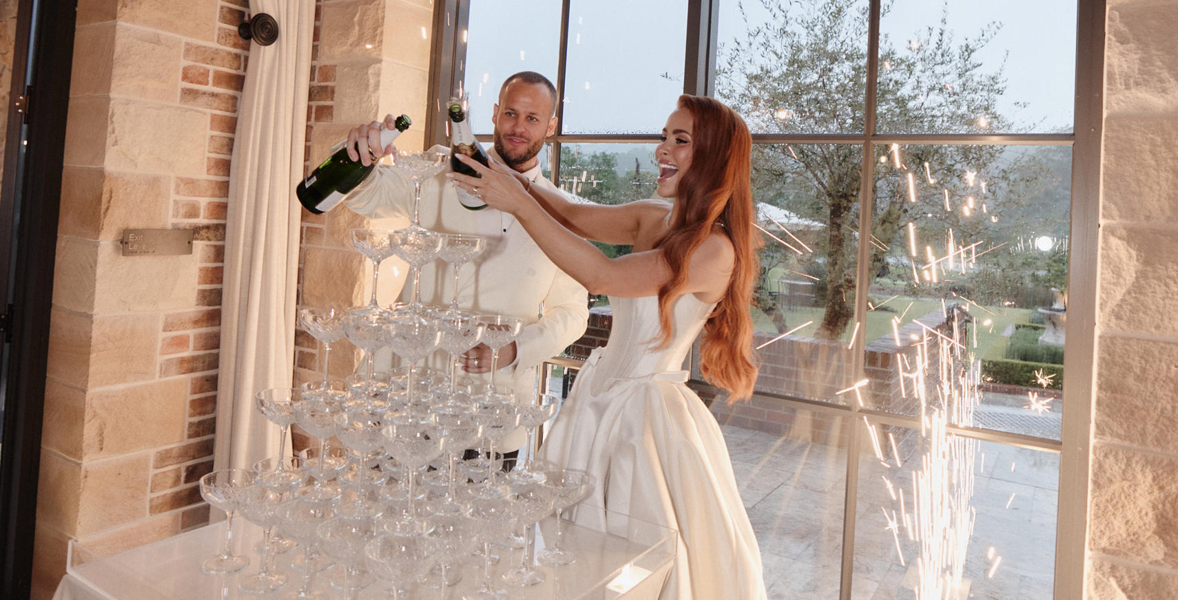 A bride and groom celebrating by pouring champagne into a pyramid of glasses, with sparklers in the background through large windows.