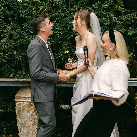 A wedding ceremony with a bride and groom holding hands, smiling at each other, with an officiant holding a book and a microphone, outdoors with greenery in the background.