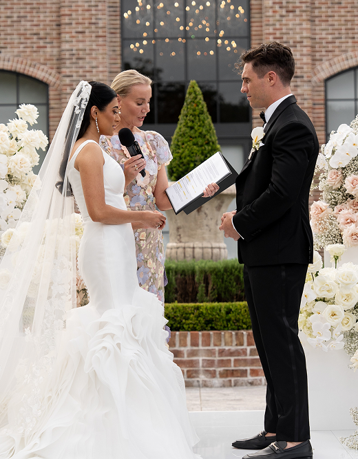Bride and groom exchanging vows during outdoor wedding ceremony with officiant, surrounded by floral arrangements.