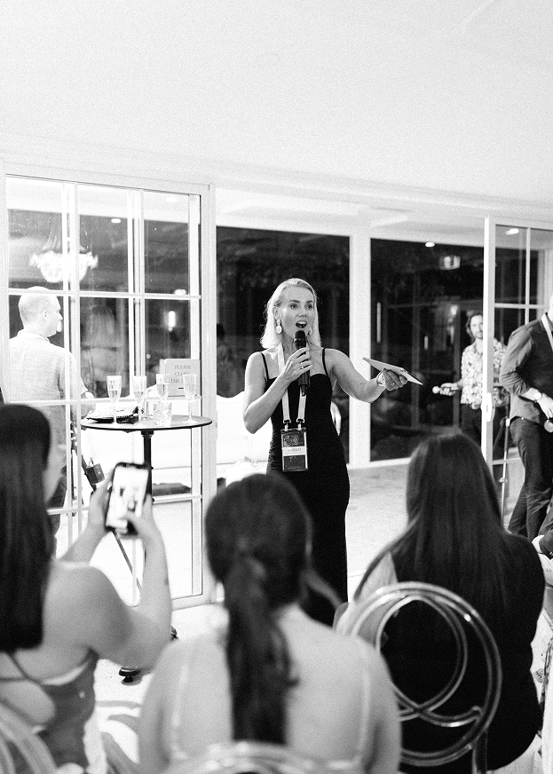 Woman speaking into a microphone at an indoor event, surrounded by seated audience members, some taking photos.