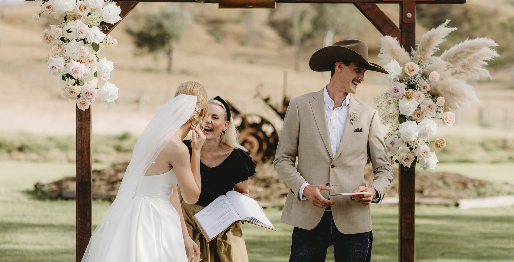 A wedding ceremony outdoors with a bride, groom, and officiant. The bride and officiant are sharing a laugh, while the groom is smiling. The ceremony features floral arrangements with white and pale pink roses and pampas grass on a wooden arch.