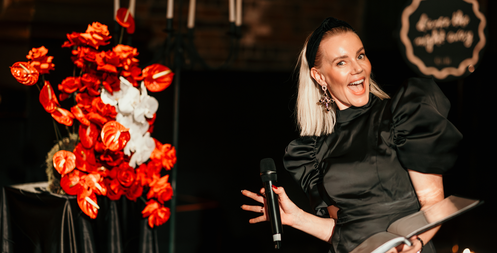A woman with blonde hair, wearing black satin puff-sleeve top and statement earrings, holding a microphone and a tablet, smiling at a party or event with a large red and white floral arrangement in the background.