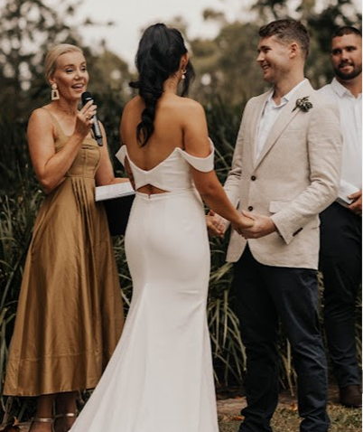 A bride and groom hold hands during their wedding ceremony outdoors, with a woman officiant speaking into a microphone nearby.