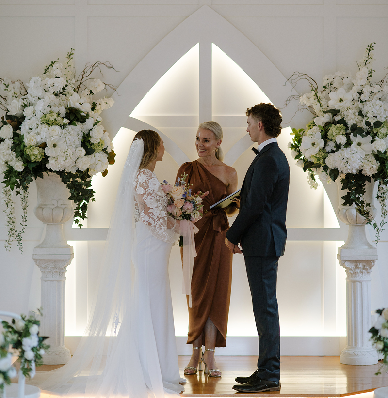 A wedding ceremony with a bride and groom facing an officiant, surrounded by large floral arrangements in white vases with white flowers and greenery, inside a decorated venue with geometric lighting and panels.