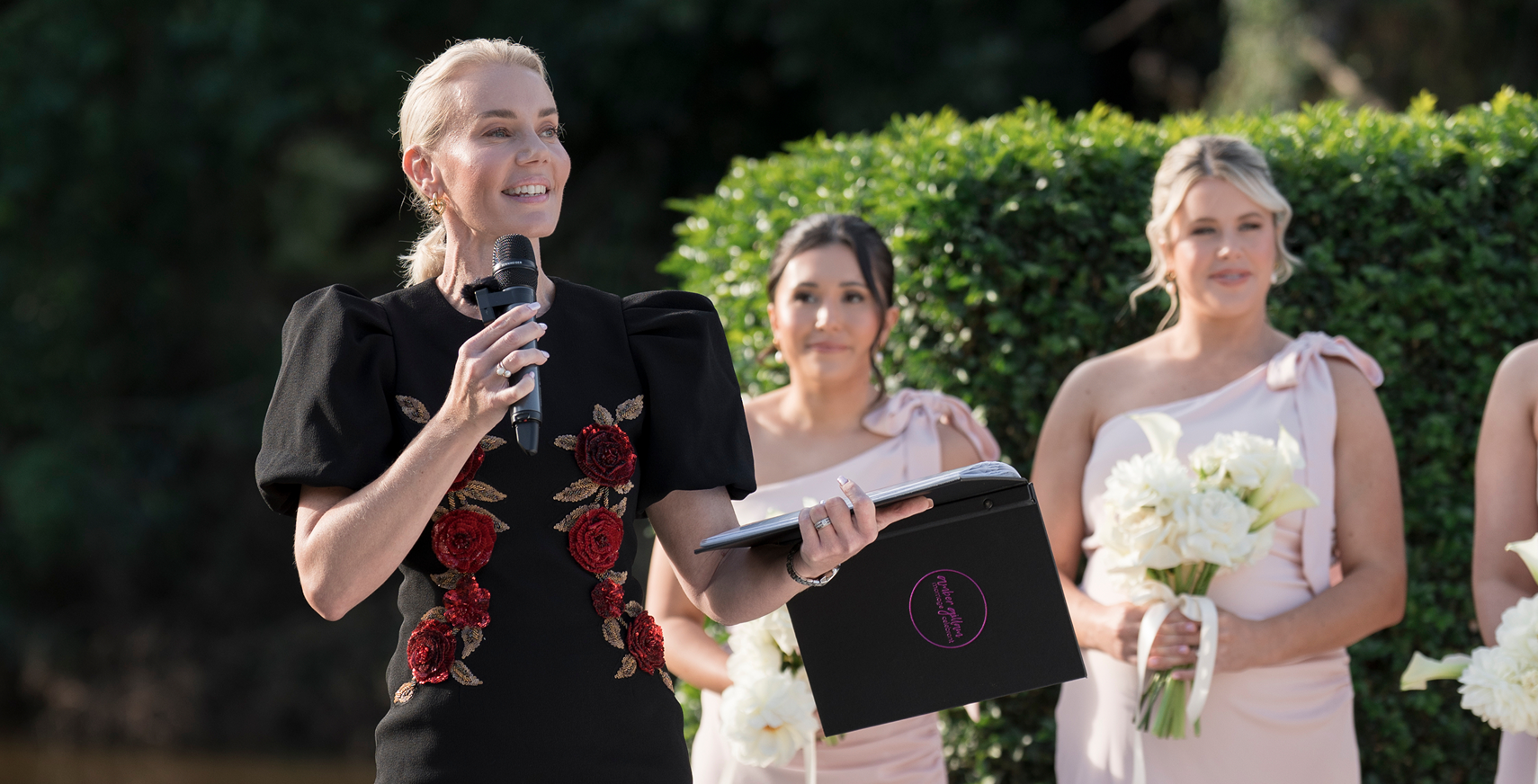 Lady in black dress giving speech at outdoor wedding ceremony, holding a microphone and a folder, with bridesmaids holding white bouquets standing behind her.