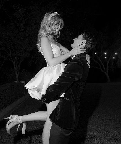 A man lifting a woman in a dress outdoors at night, both smiling and looking at each other.