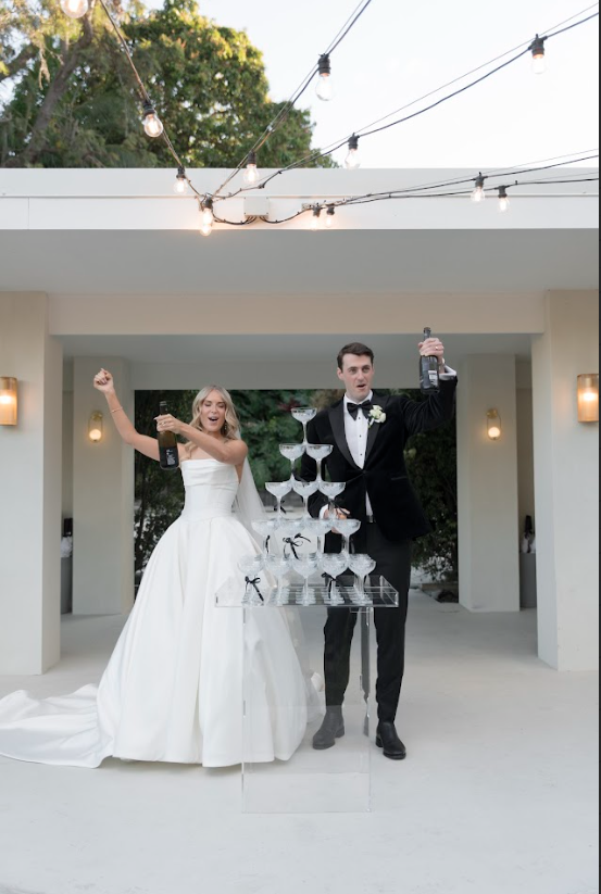 Bride and groom celebrating at their wedding in an outdoor venue with string lights, holding champagne bottles, with a tower of stacked glasses on a clear table.