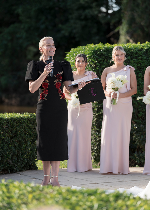 A woman in a black dress with red floral embroidery is speaking into a microphone at an outdoor wedding ceremony, with bridesmaids holding bouquets in the background.