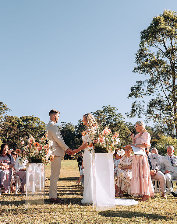 Outdoor wedding ceremony with a couple holding hands, surrounded by seated guests, large floral arrangements, and a woman officiating with a microphone, under a clear blue sky.