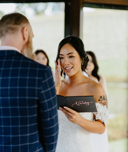 Bride smiling and holding a small black book during her wedding ceremony, with a man in a blue checkered suit facing her.