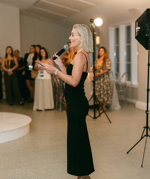 Woman in black dress speaking into a microphone at an indoor event, with an audience and photography equipment in the background.