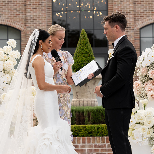 A bride and groom attend a wedding ceremony outdoors, with the officiant reading from a book. The bride is dressed in a white wedding gown with a veil, holding a microphone. The groom is dressed in a black tuxedo. Floral arrangements and a brick building are in the background.