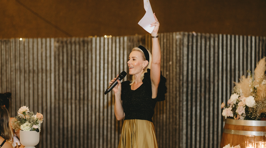 A woman with blonde hair, wearing a black top and a gold skirt, holding a microphone in one hand and a paper in the other, speaking at an event with floral decorations.