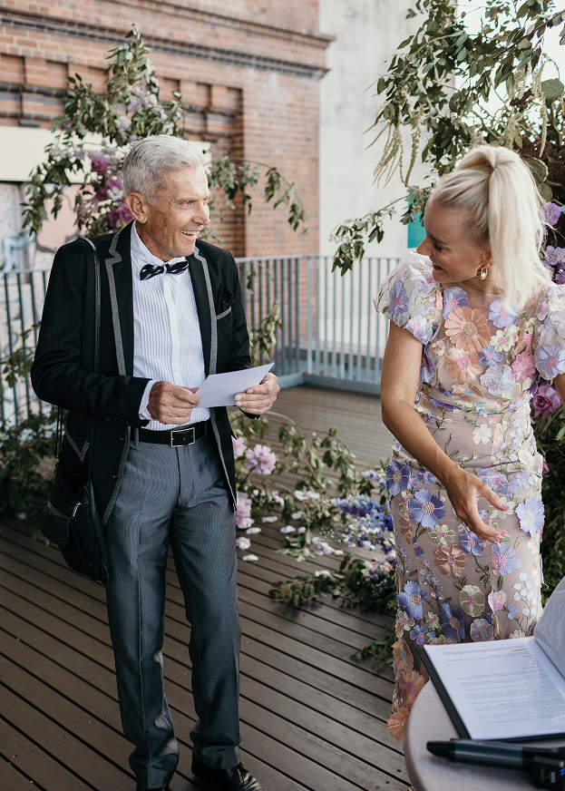 A joyful elderly man in a tuxedo and a stylish young woman in a floral dress at a wedding or celebration reception, sharing a happy moment on a decorated outdoor terrace.