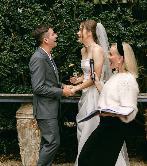 A bride and groom exchanging rings during their wedding ceremony outdoors, with an officiant holding a microphone and a book, all smiling and facing each other.