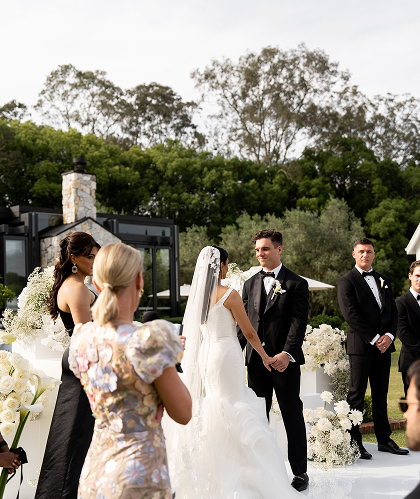 A wedding ceremony outdoors with a bride and groom holding hands, surrounded by family and friends dressed in formal attire, with a decorated aisle and lush greenery in the background.